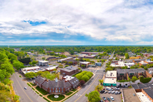 An aerial view of a small town with brick buildings, green trees, and paved roads under a partly cloudy sky.