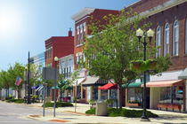 A sunlit street scene in a historic downtown with brick buildings, storefronts with awnings, and trees along the sidewalk.