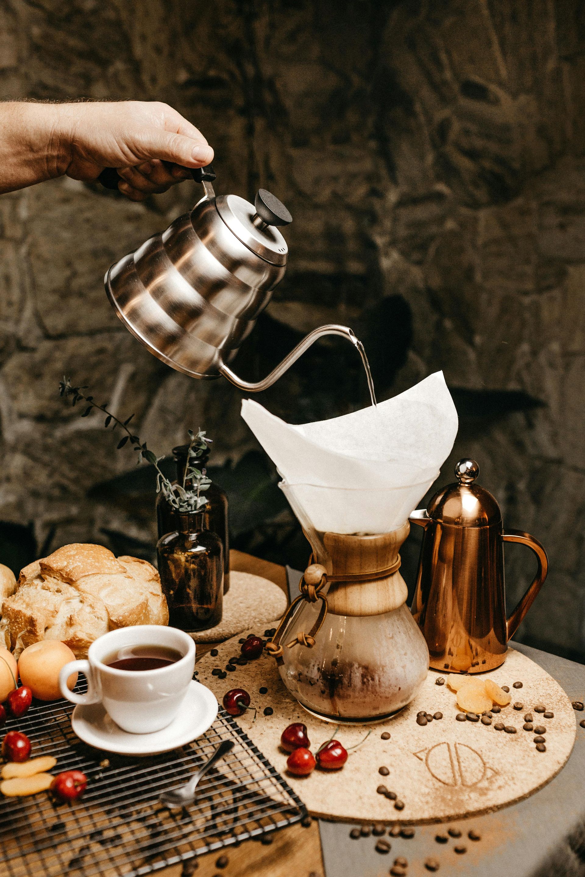 Hand pouring water into a pour-over coffee maker. Coffee setup includes coffee cup, bread, fruit, and gold kettle on a table.
