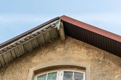 A weathered beige stucco gable wall under a blue sky, featuring a damaged wooden eave and dark brown roof trim.