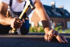 A roofer holds a hammer above a nail to secure black shingles on a house roof.