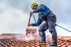 A person wearing safety gear and a harness pressure-washes a red tiled roof while tethered to a safety rope.