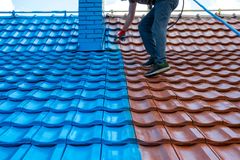 A person uses a paint sprayer to coat half of a tiled roof in bright blue, contrasting with the remaining red tiles.