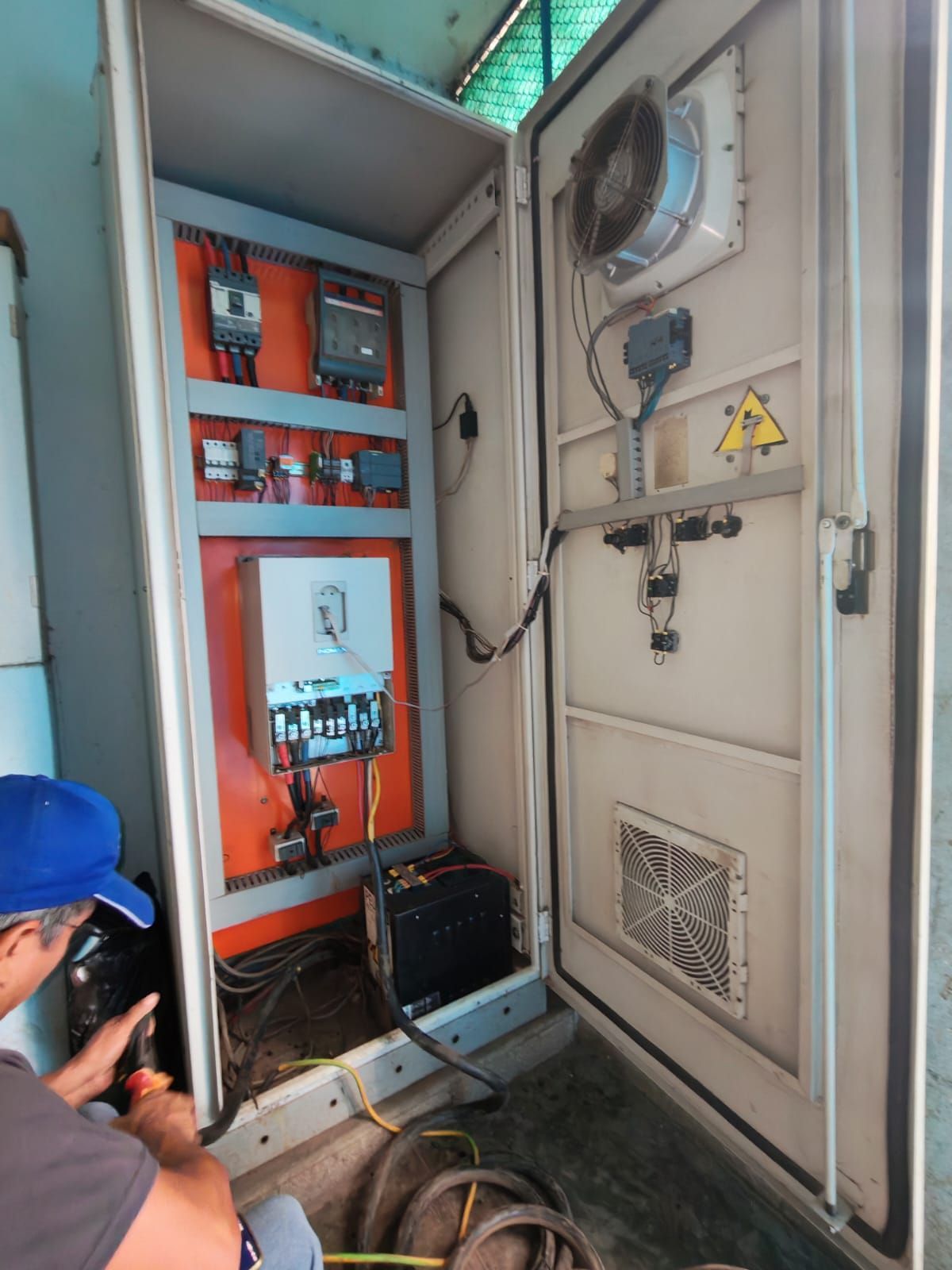 Armario de control eléctrico abierto con cableado, señal de advertencia y un trabajador inspeccionándolo.