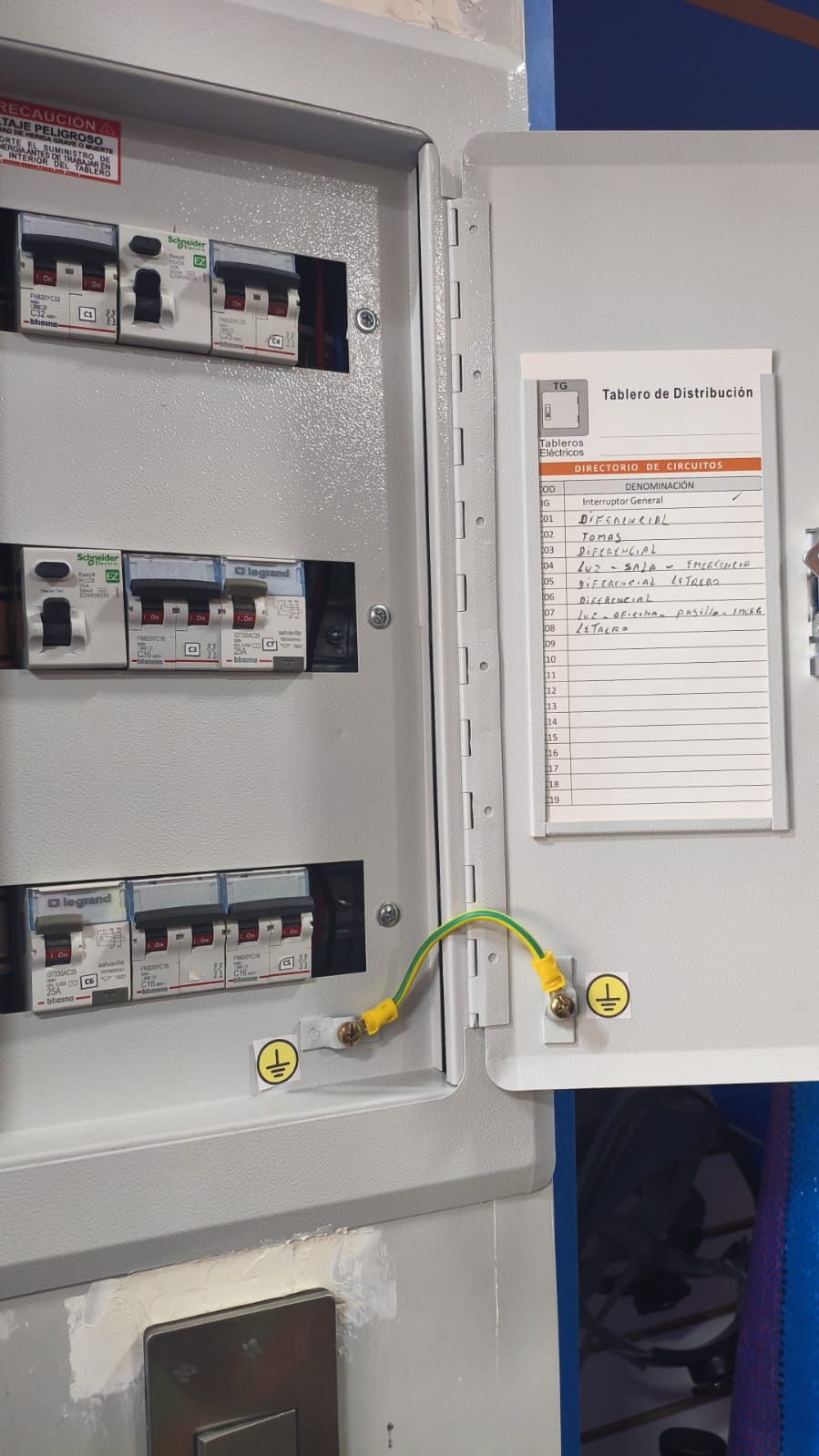 Panel de disyuntores eléctricos abierto con circuitos etiquetados y precintos de seguridad amarillos en la puerta.