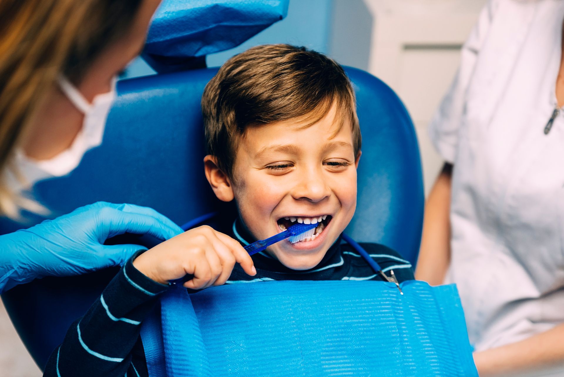 A young boy sits in a dental chair while a dentist shows him how to brush his teeth