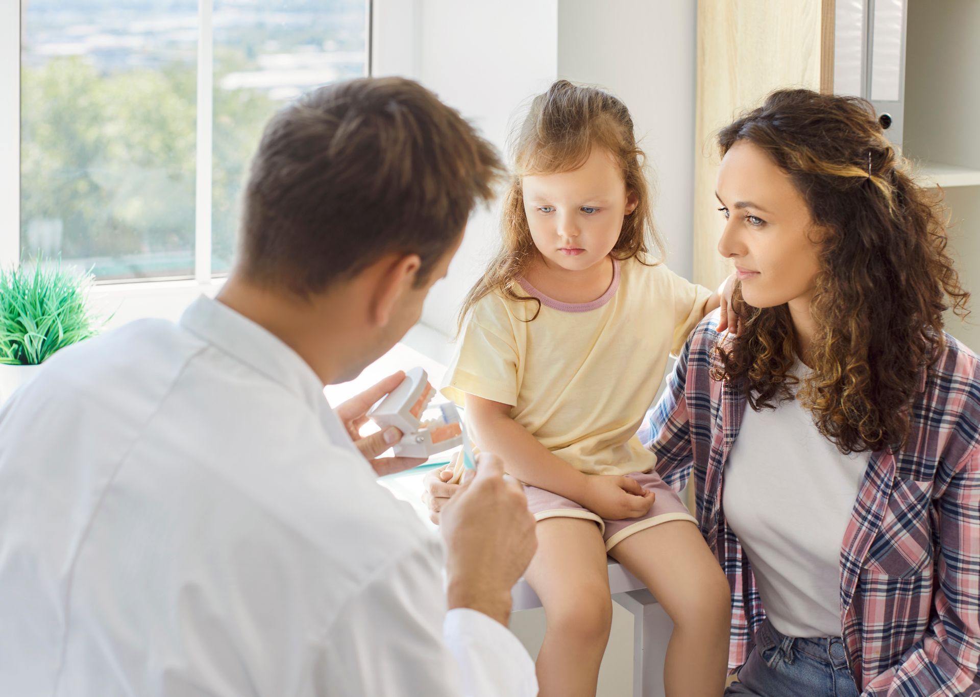 A dentist shows a teeth model to a young girl and her mother in a dental office