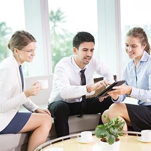 Three colleagues meeting on a sofa, reviewing a tablet with coffee cups on the table.