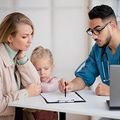 Doctor showing a mother and child a medical form at a desk, with a clipboard and laptop nearby.
