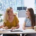 Two coworkers sit at a table, looking at each other in a bright office meeting room.