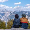 Two people sit on a rocky overlook, facing a snowy mountain range under a blue sky.