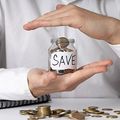 Hands sheltering a jar labeled “SAVE” above scattered coins on a table