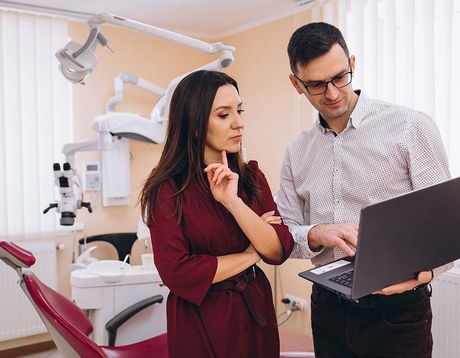 Two people discussing a laptop in a dental clinic with an exam chair and medical equipment in the background