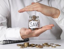 Hands holding a jar labeled “SAVE” above scattered coins and bills on a white table