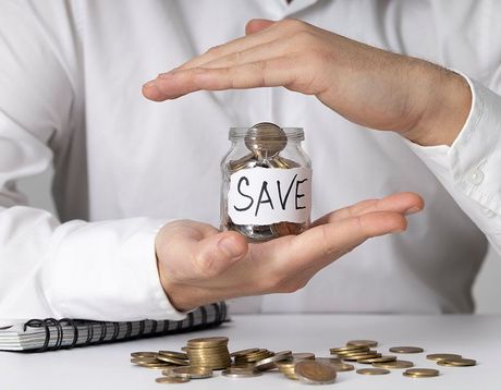 Hands holding a jar labeled “SAVE” above scattered coins and bills on a white table