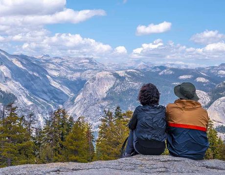 Two hikers sit on a rock overlooking a snowy mountain valley under a cloudy blue sky.
