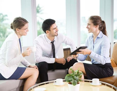 Three coworkers talking on a couch in a bright office lounge, holding coffee and a tablet.