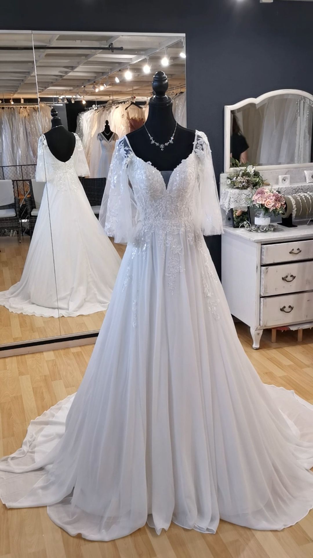 Weißes Brautkleid mit Spitzenärmeln und fließendem Rock, ausgestellt an einer Schaufensterpuppe in einem Brautmodengeschäft