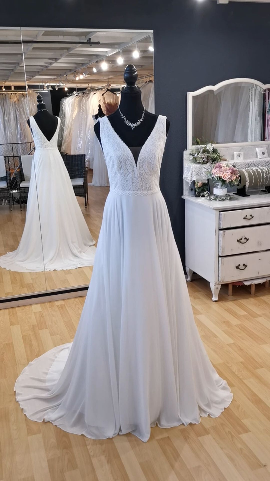 Ein weißes Brautkleid aus Spitze wird auf einer Schaufensterpuppe in einem Brautmodengeschäft ausgestellt; im Hintergrund sind Spiegel und ein Schminktisch zu sehen.