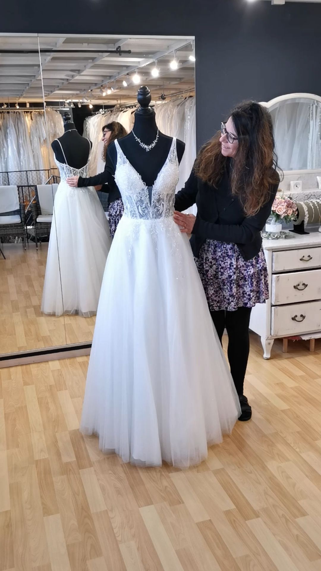 Brautmodengeschäft mit einem weißen Brautkleid an einer Schaufensterpuppe, eine Frau, die es vor einem Spiegel zurechtzupft.