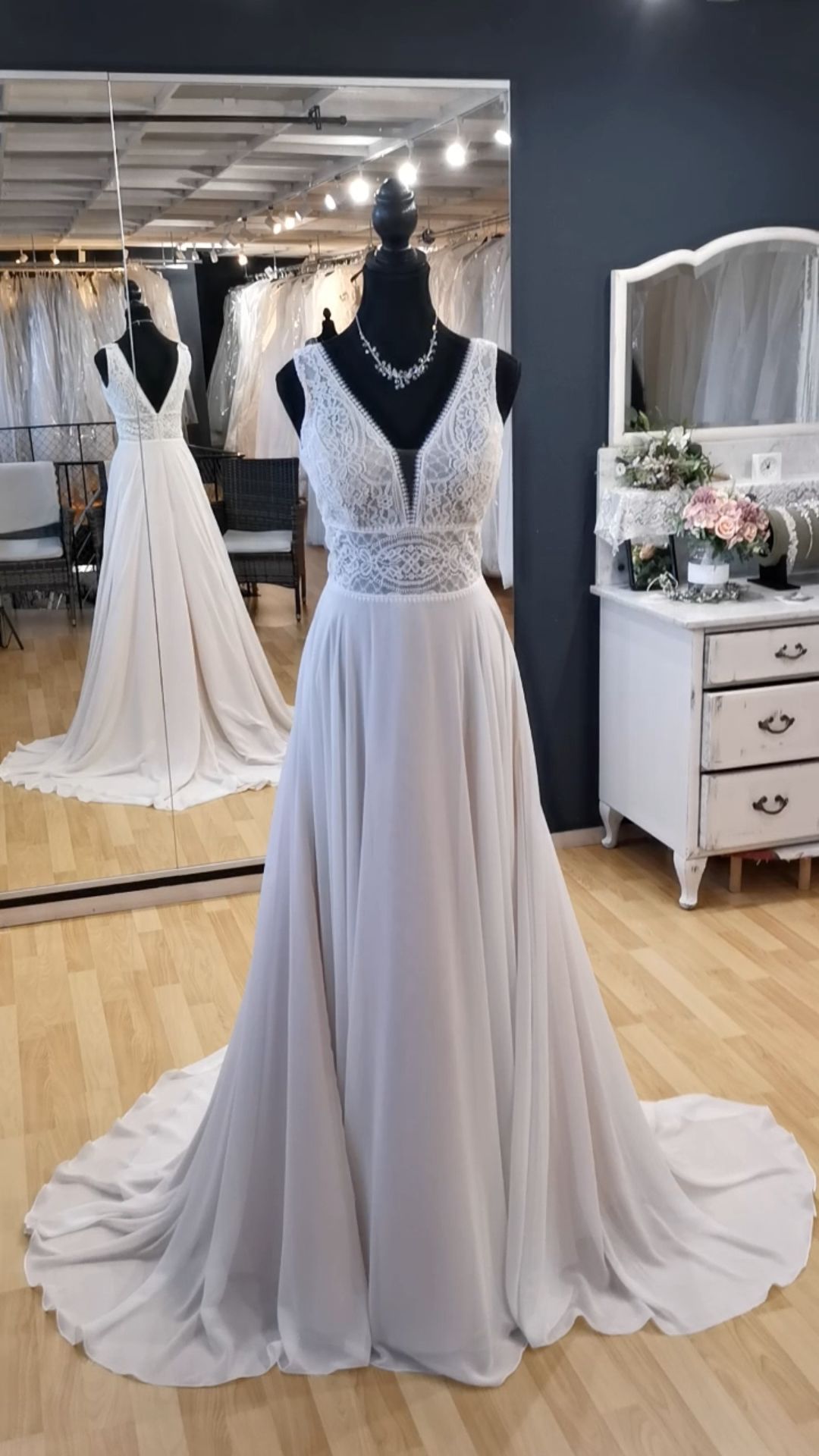Weißes Brautkleid aus Spitze an einer Schaufensterpuppe in einem Brautmodengeschäft, mit fließender Schleppe und Spiegel im Hintergrund.
