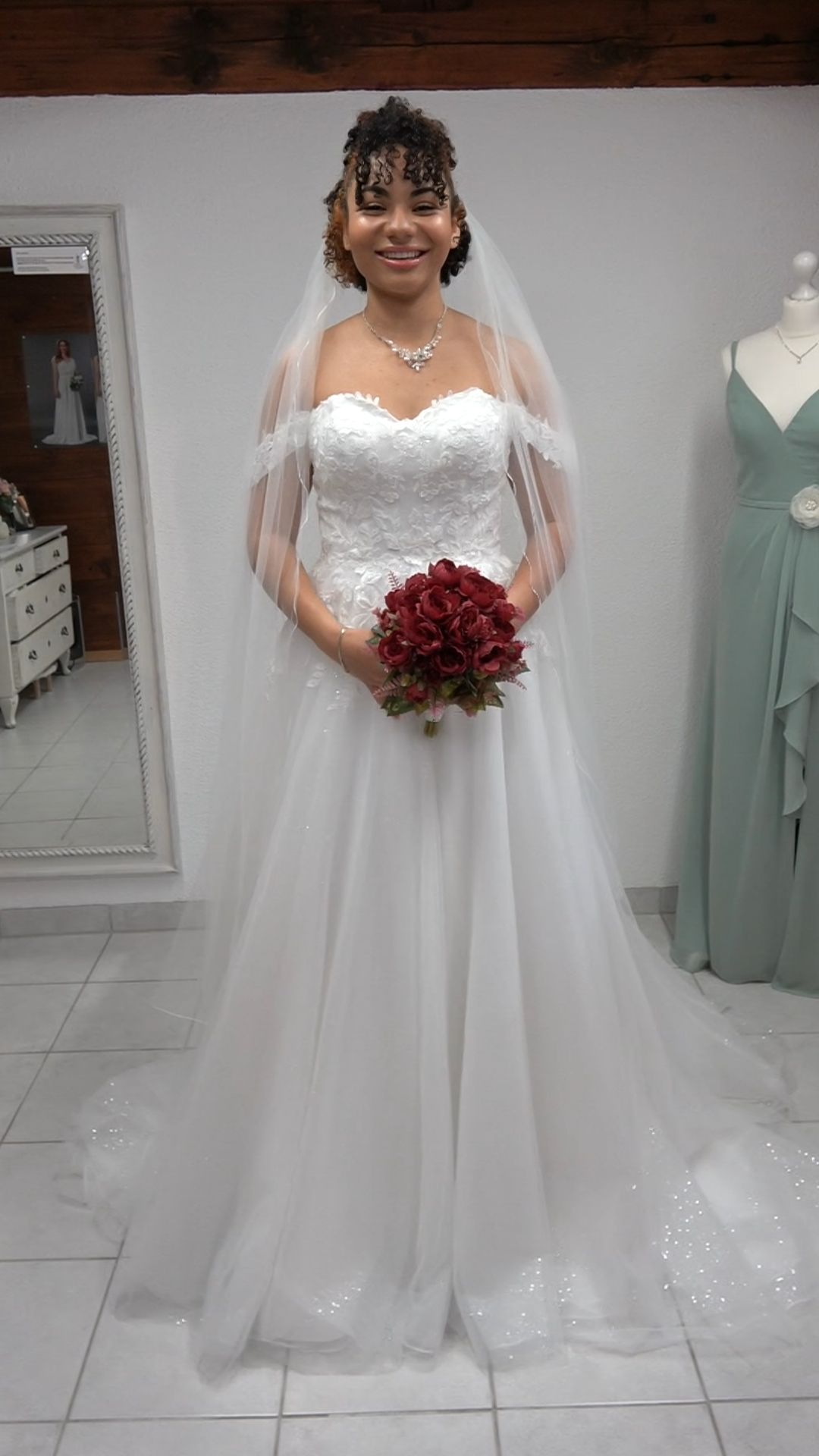 Eine Braut in einem weißen Hochzeitskleid und Schleier hält einen roten Brautstrauß und steht in einem Brautmodengeschäft.