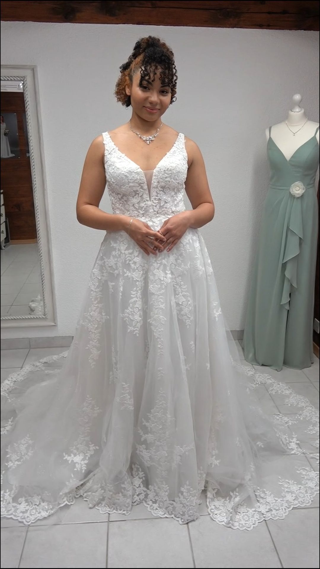 Frau in einem weißen Spitzenhochzeitskleid steht in einer Brautboutique neben einer mintgrünen Kleiderpuppe