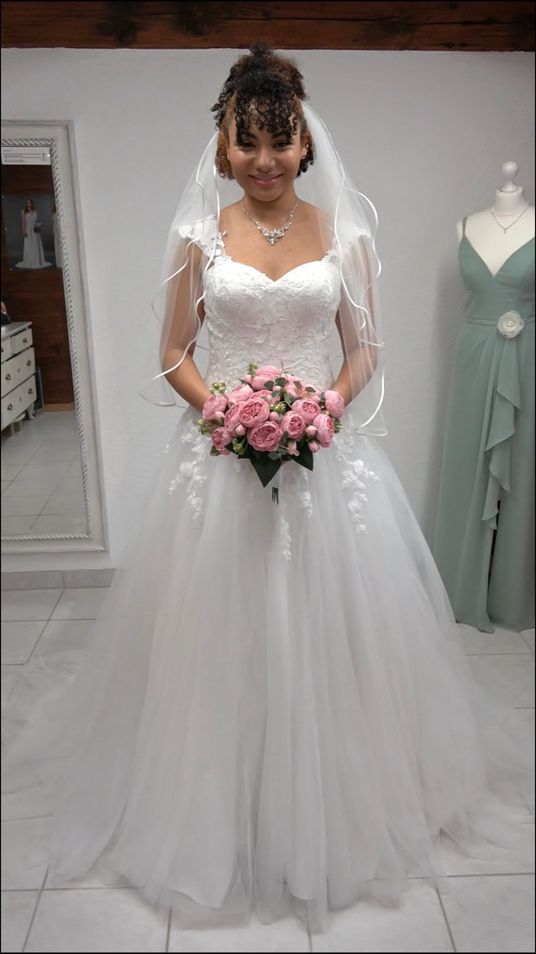 Braut in einem weißen Hochzeitskleid, die in einem Brautgeschäft einen rosa Brautstrauß hält und unter einem Schleier lächelt