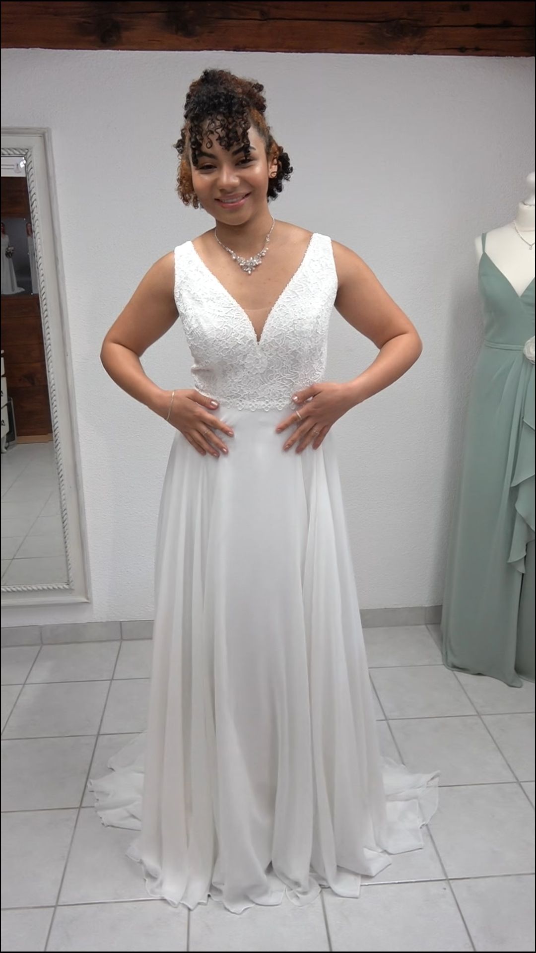 Frau in einem weißen Brautkleid steht lächelnd in einer Boutique, die Hände in die Hüften gestemmt.