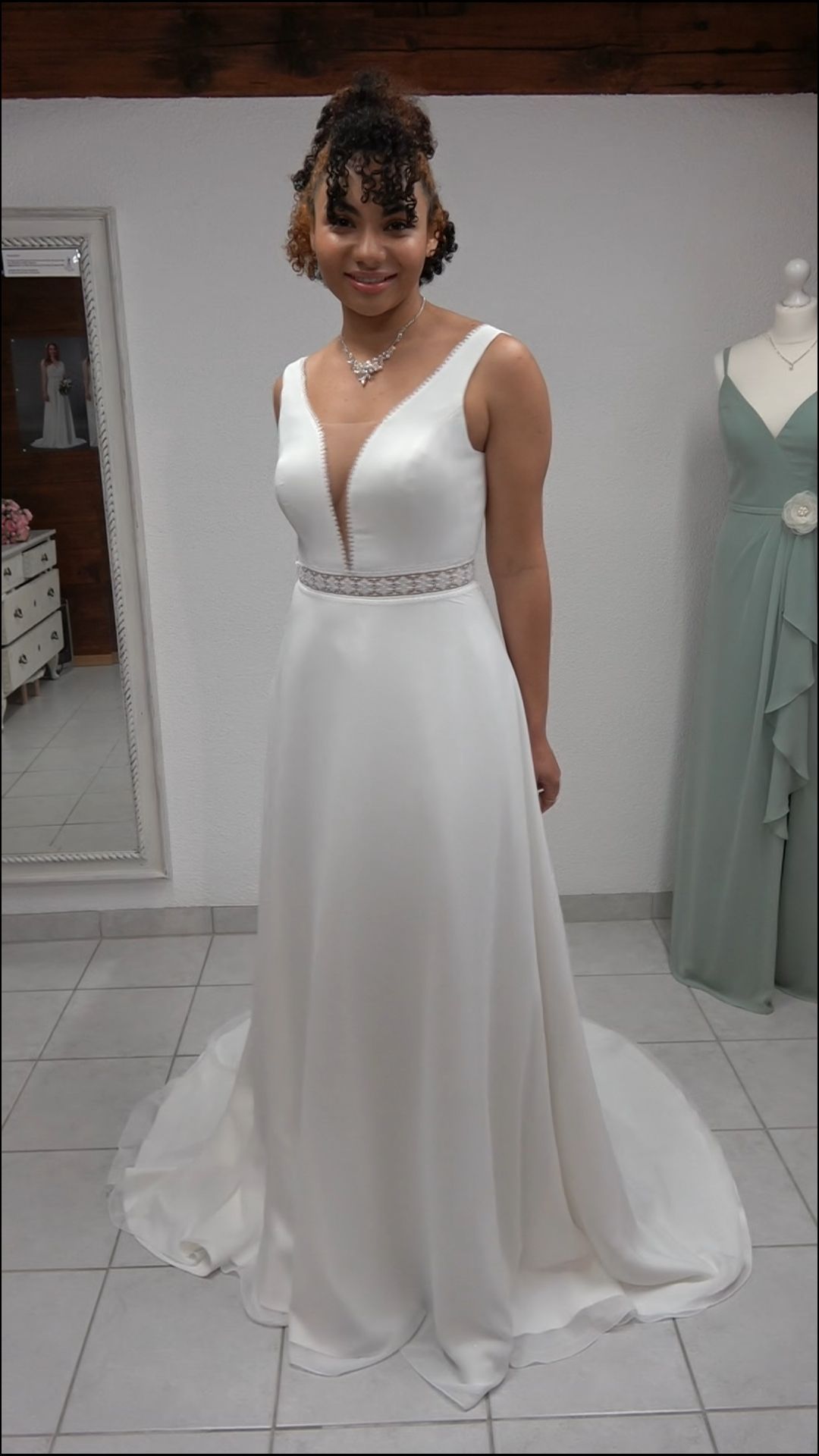 Frau in einem weißen, ärmellosen Kleid mit fließender Schleppe steht in einem Innenraum auf einem gefliesten Boden.