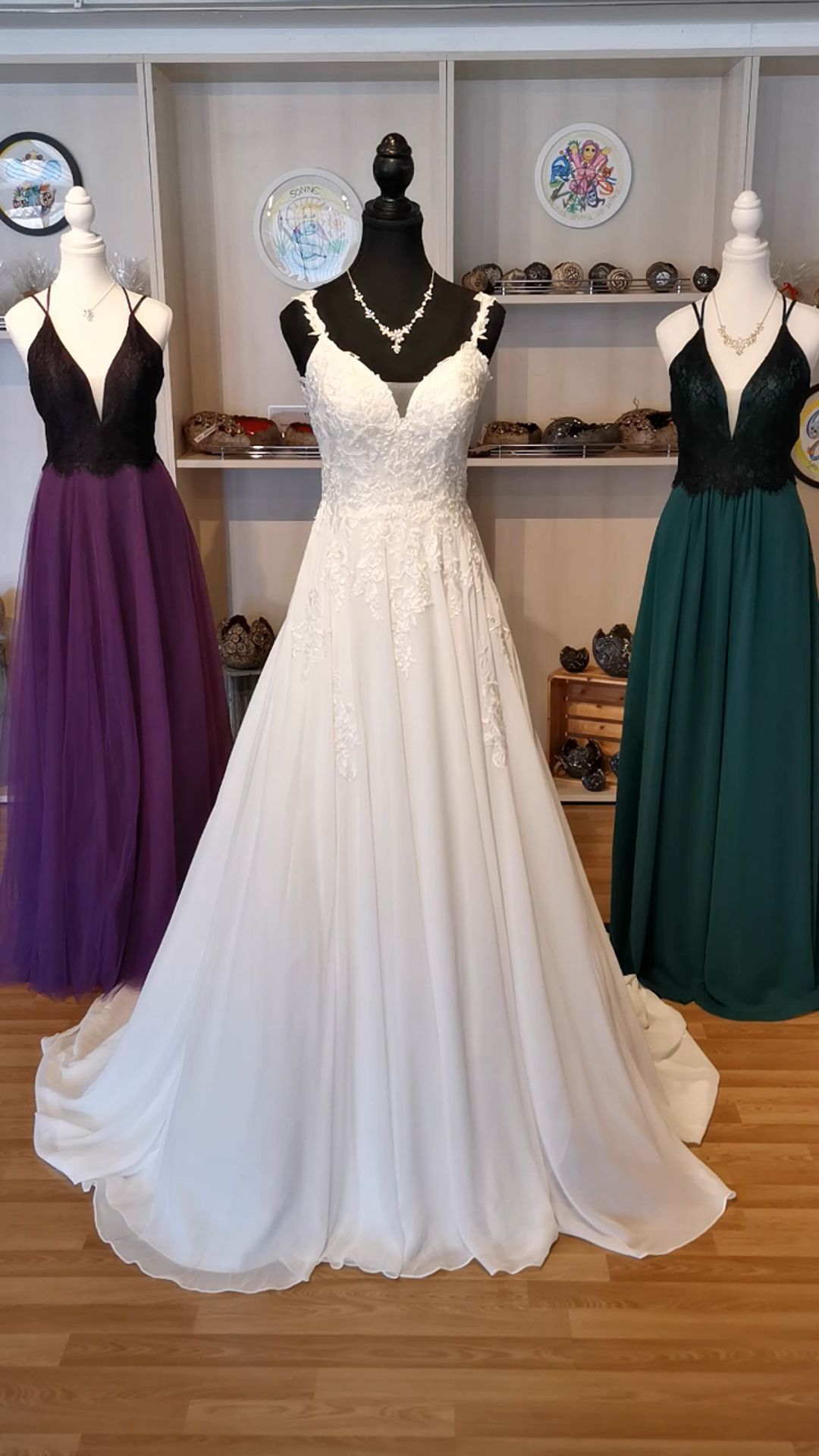Weißes Spitzenhochzeitskleid an einer Schaufensterpuppe, flankiert von lila und grünen Brautjungfernkleidern in einer Boutique.