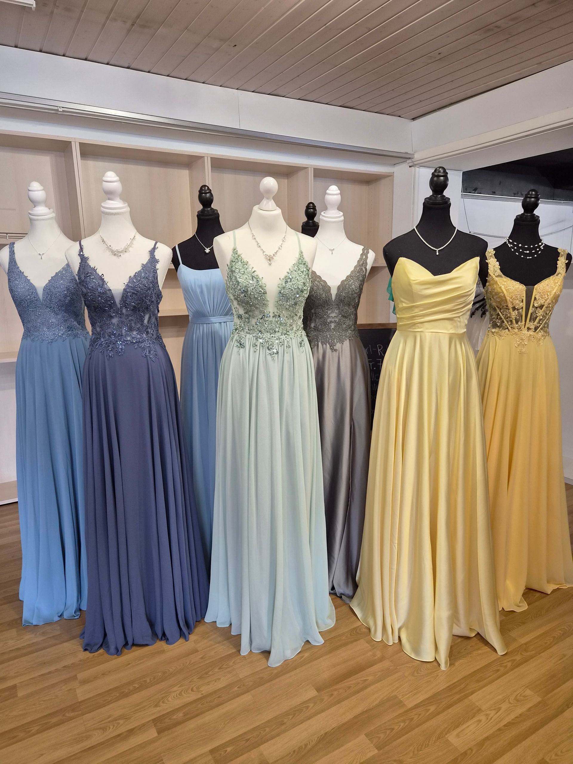 Schaufensterpuppen präsentieren elegante Abendkleider in Blau, Mint, Schwarz und Gelb in einem Brautmodengeschäft.