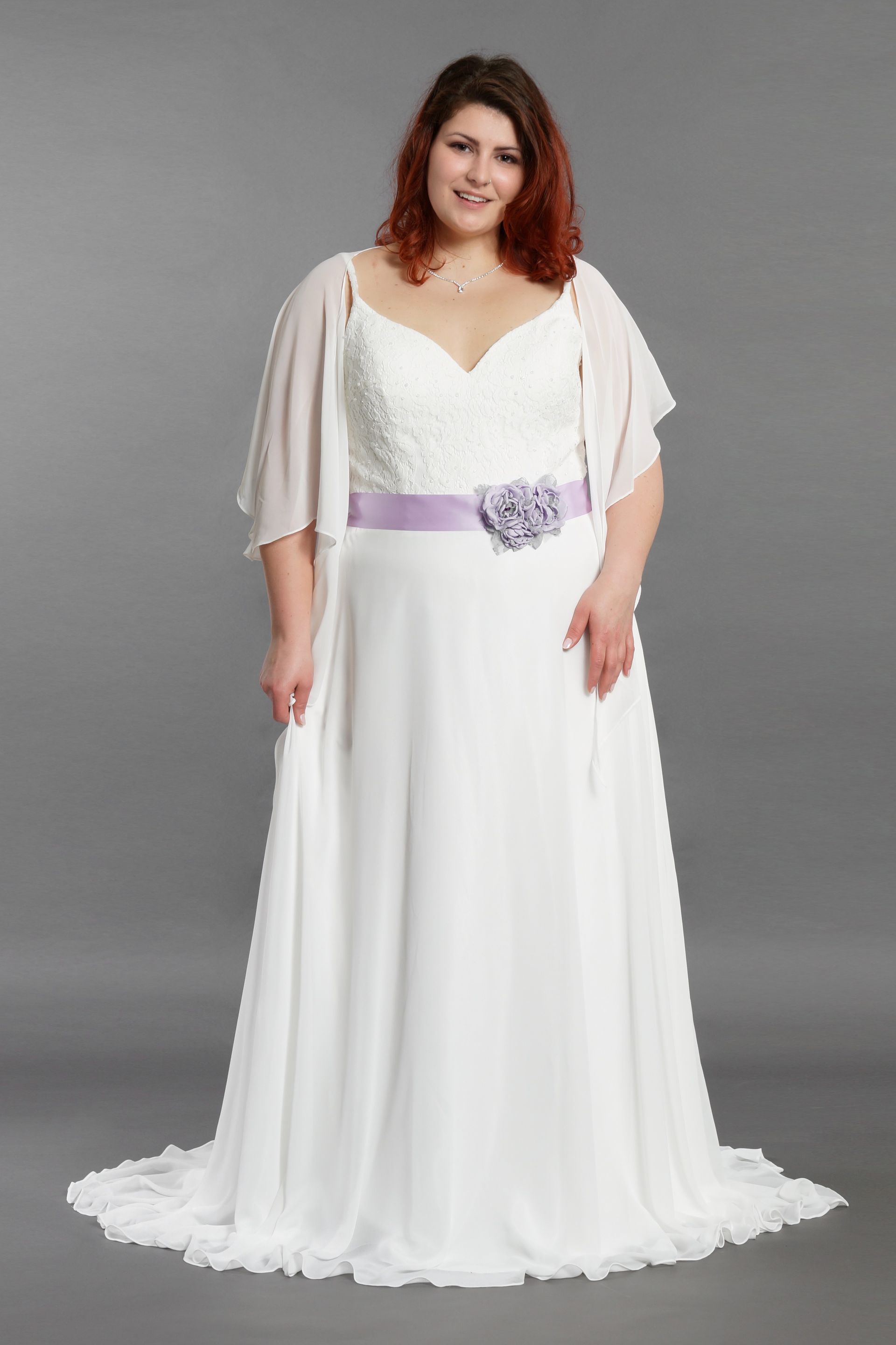 Frau in einem weißen, bodenlangen Kleid mit lavendelfarbener Schärpe, vor grauem Hintergrund.