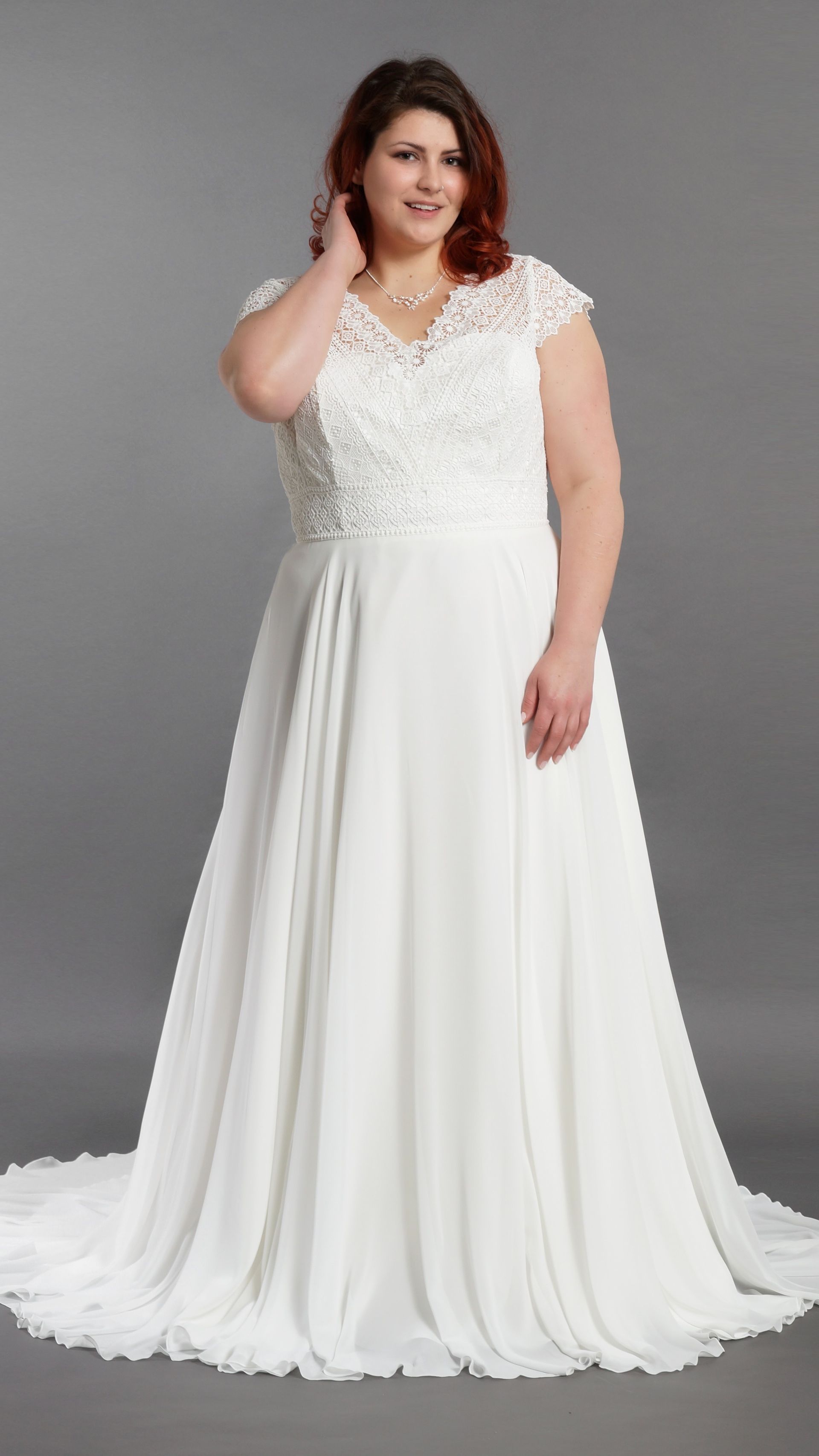 Frau in einem weißen Spitzenhochzeitskleid vor grauem Hintergrund, Hand nahe dem Gesicht
