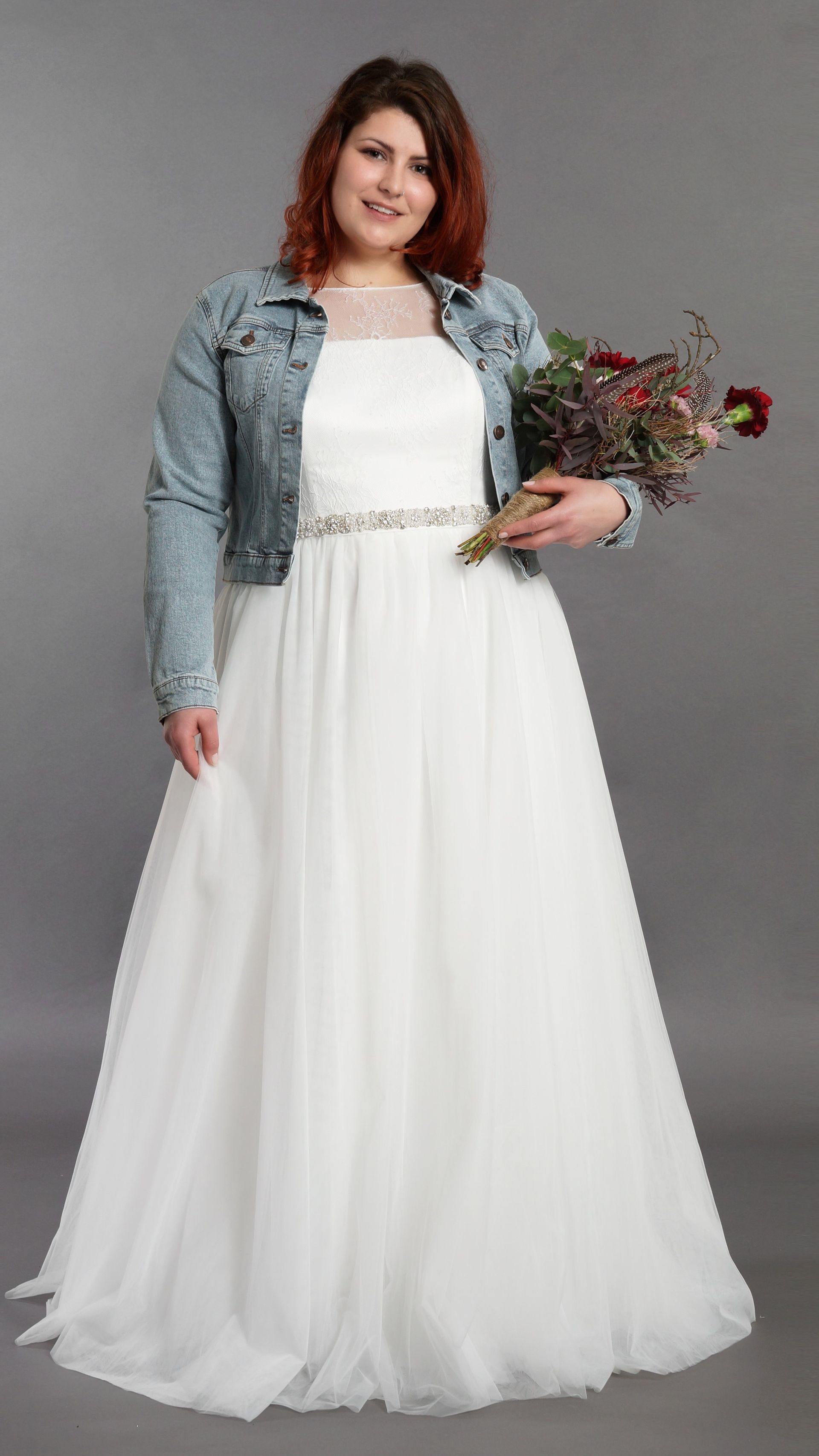 Frau in weißem Kleid und Jeansjacke hält einen Blumenstrauß vor grauem Hintergrund