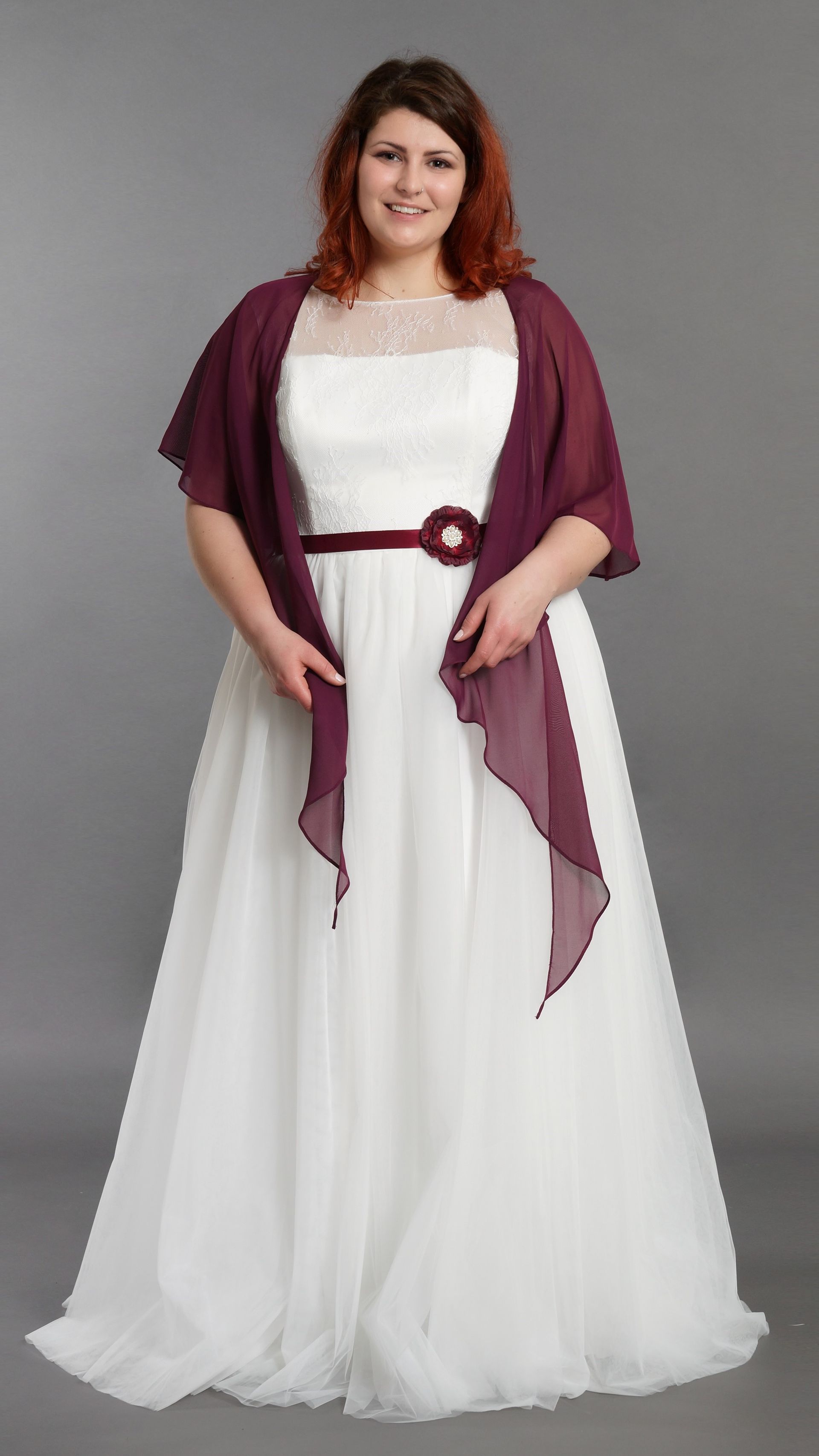 Frau in einem weißen, bodenlangen Kleid mit bordeauxrotem Schal und Gürtel, vor einem grauen Hintergrund.