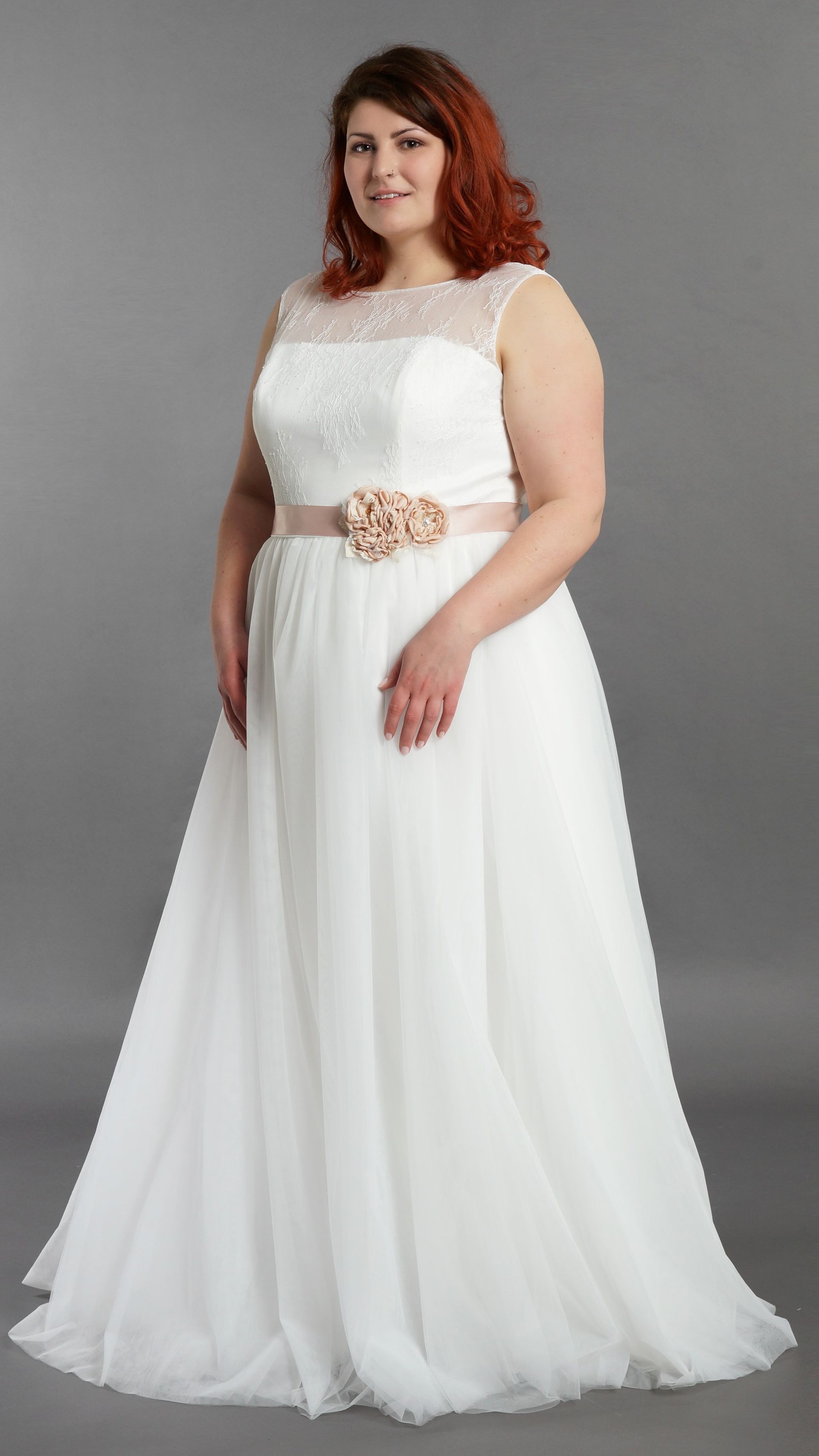 Frau in einem weißen, ärmellosen, bodenlangen Kleid mit Perlengürtel, vor grauem Hintergrund.