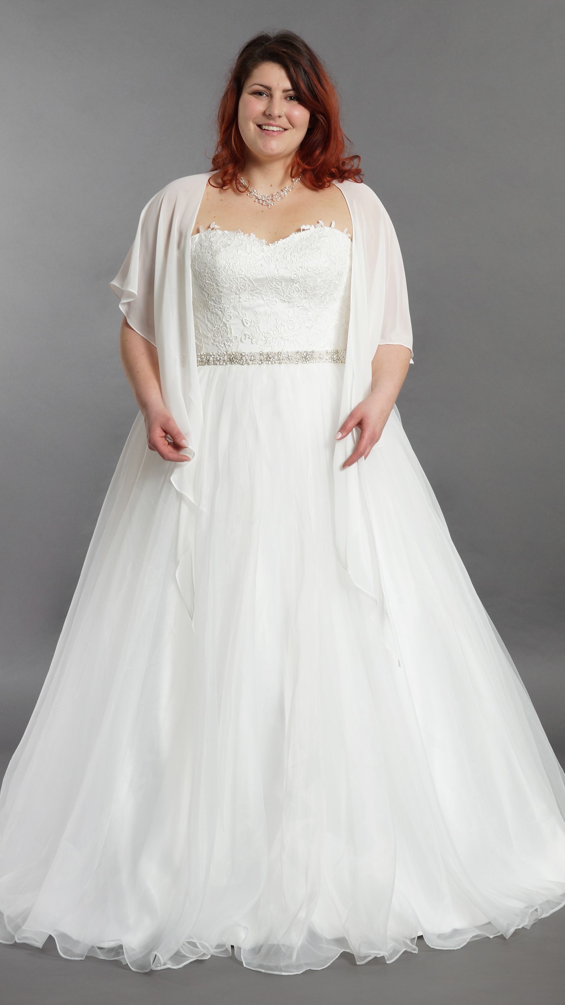 Eine Frau präsentiert vor grauem Hintergrund ein weißes, bodenlanges Kleid mit paillettenbesetztem Oberteil und transparenten Cape-Ärmeln.