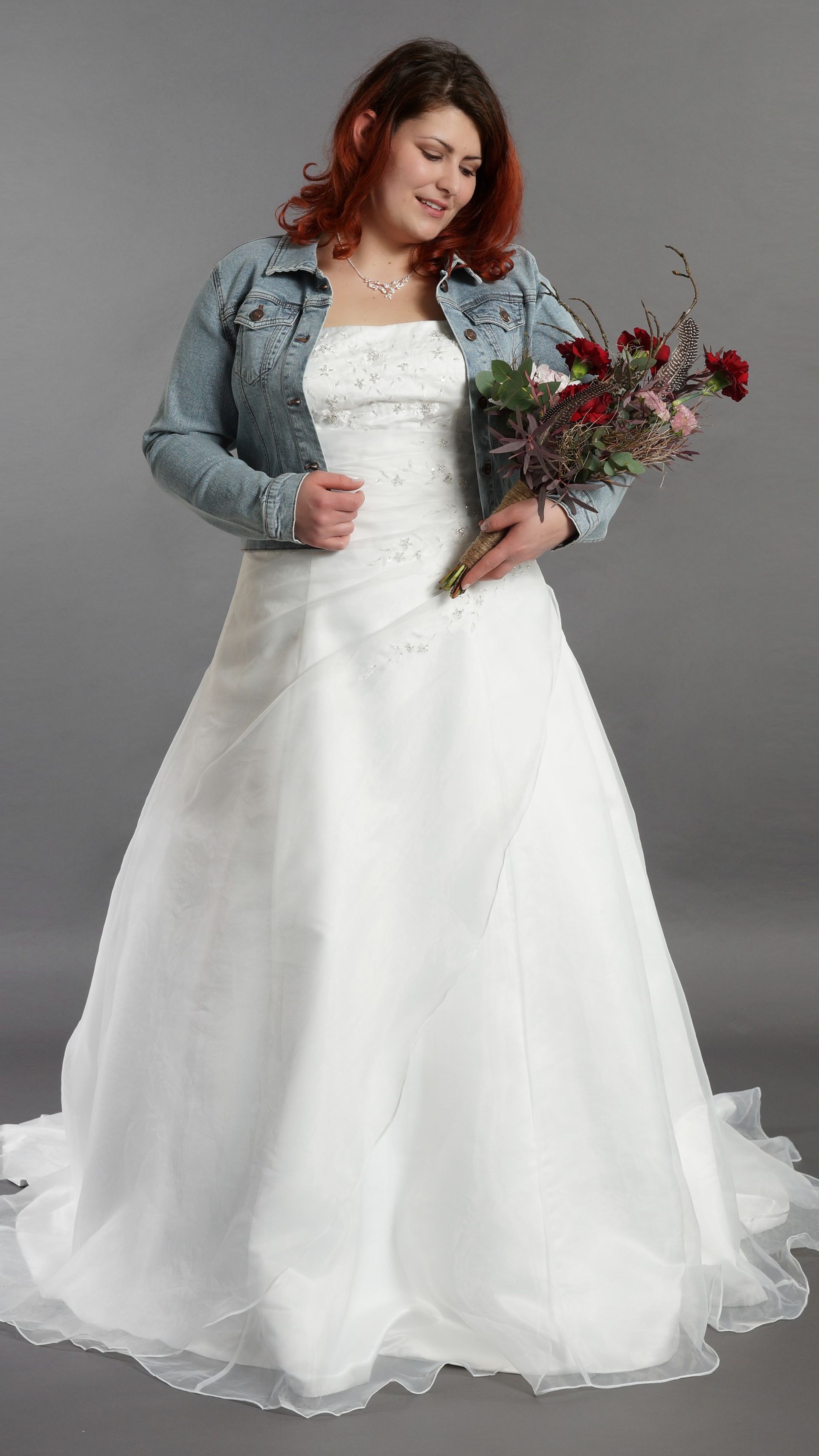 Braut in weißem Kleid und Jeansjacke hält einen Brautstrauß vor grauem Hintergrund