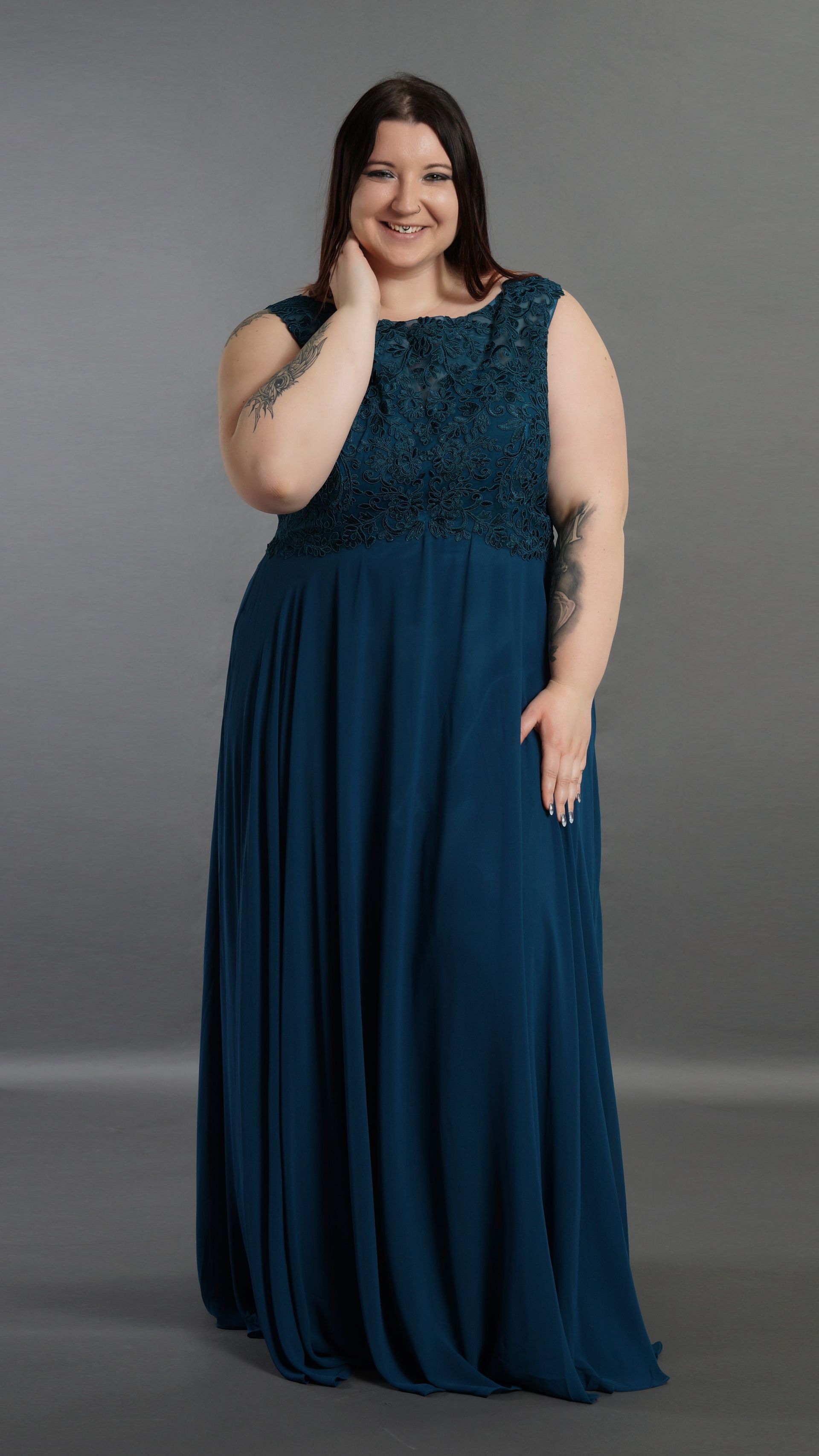 Frau in einem marineblauen, bodenlangen Kleid vor grauem Hintergrund, lächelnd mit einer Hand im Haar.