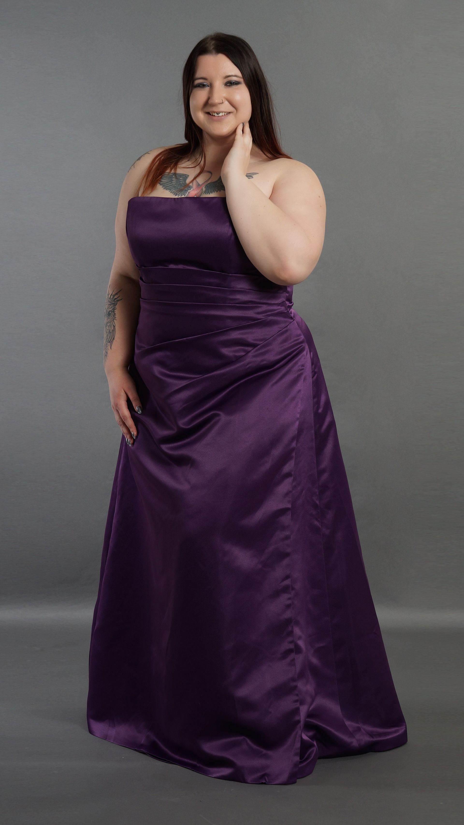 Frau in einem violetten Satinkleid posiert vor einem grauen Hintergrund, lächelt und hält das Kleid fest.