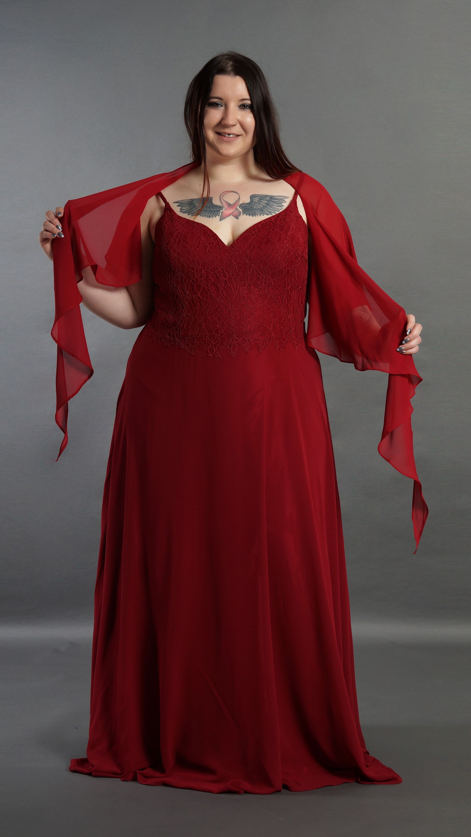Frau in einem tiefroten Kleid mit transparenten Ärmeln, vor einem grauen Studiohintergrund.