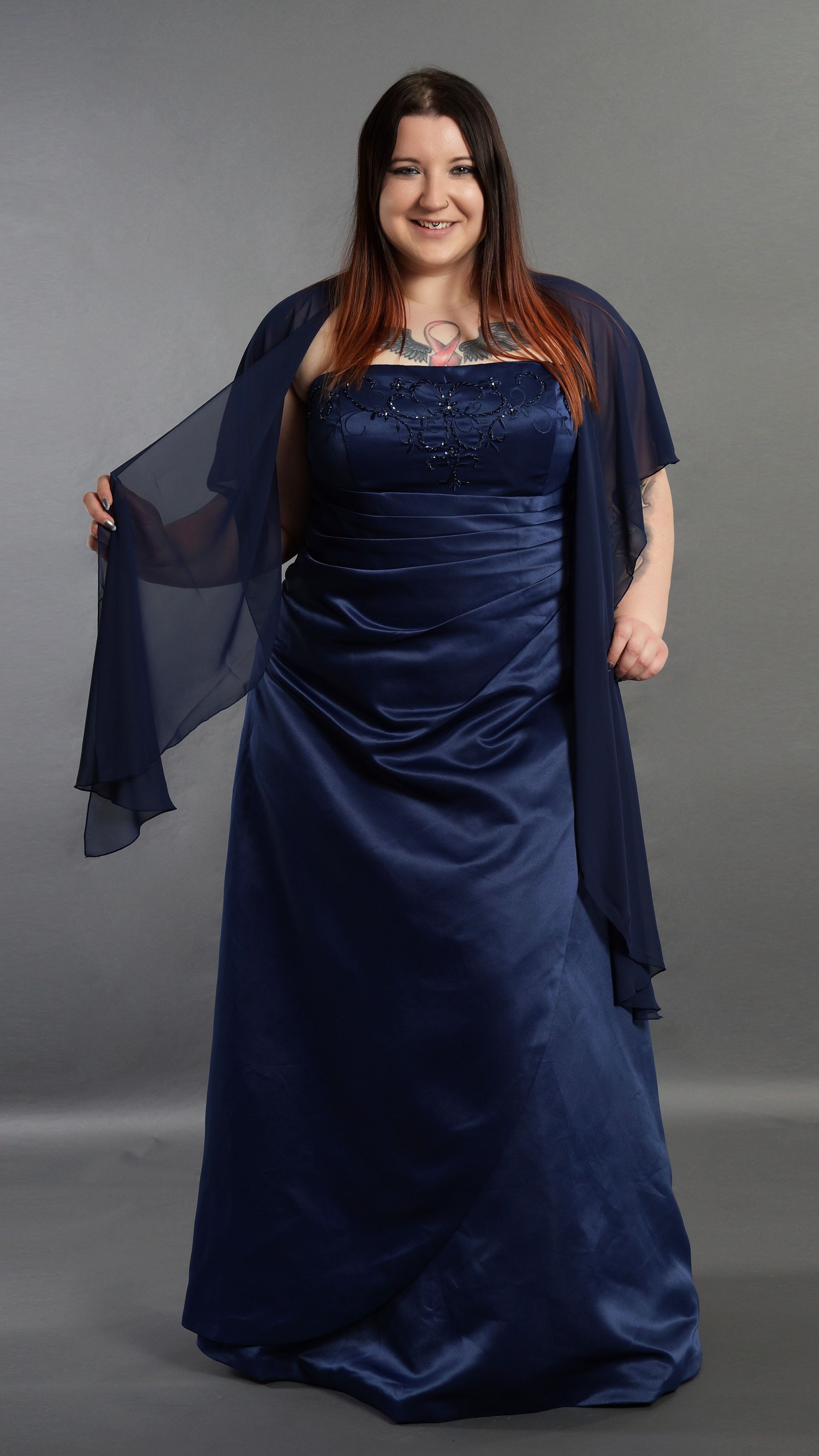 Eine Frau in einem marineblauen Abendkleid steht vor einem grauen Hintergrund und lächelt; die transparenten Ärmel fallen ihr über die Seiten.