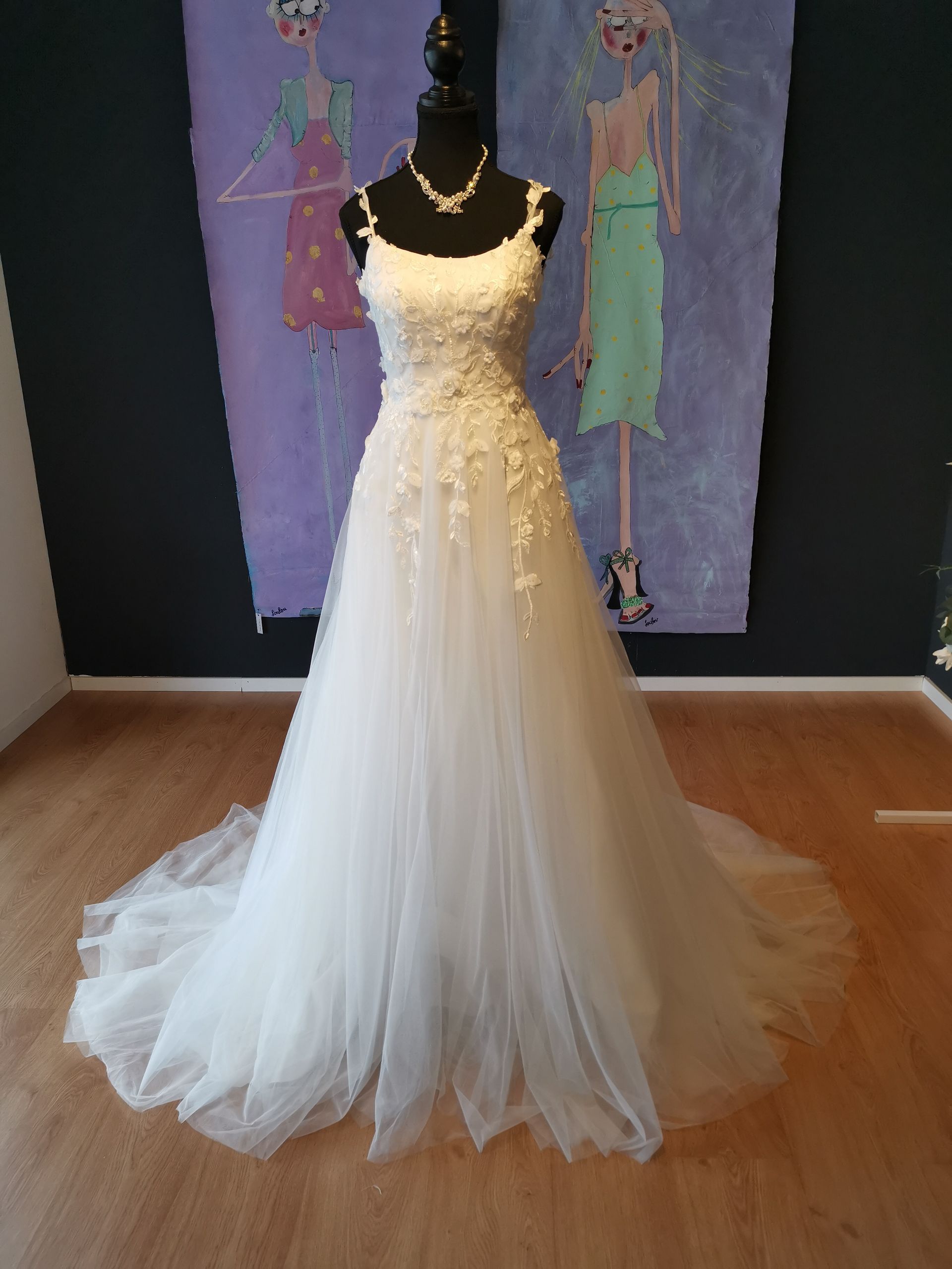 Weißes Brautkleid an einer Schaufensterpuppe, mit eng anliegendem Spitzenoberteil und fließendem Tüllrock, in einem Raum mit Kunstwerken.