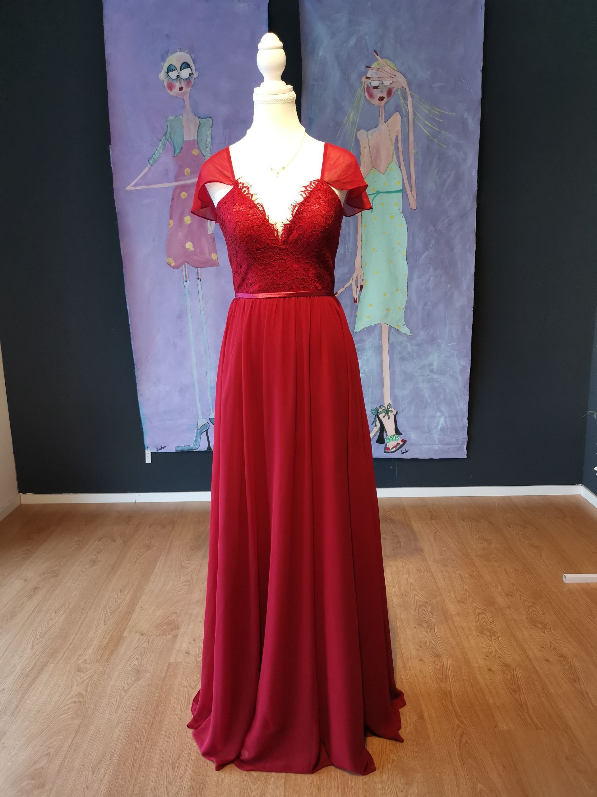 Rotes, bodenlanges Kleid an einer Schaufensterpuppe in einer Galerieumgebung, mit dunklem Hintergrund und gemalten Figuren dahinter.