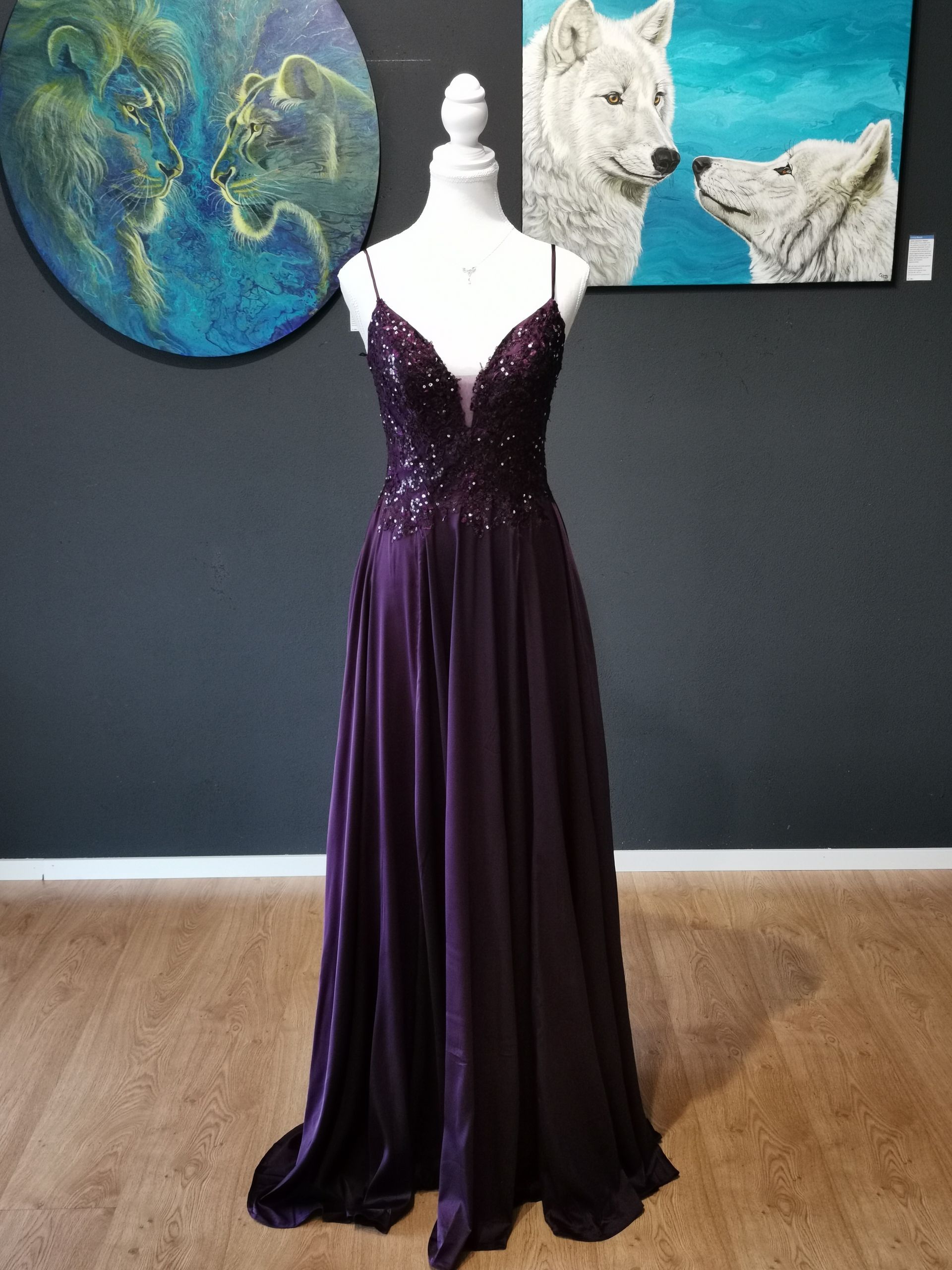 Ein tiefviolettes, paillettenbesetztes Abendkleid an einer Schaufensterpuppe in einem galerieartigen Raum mit Kunstwerken an der Wand.