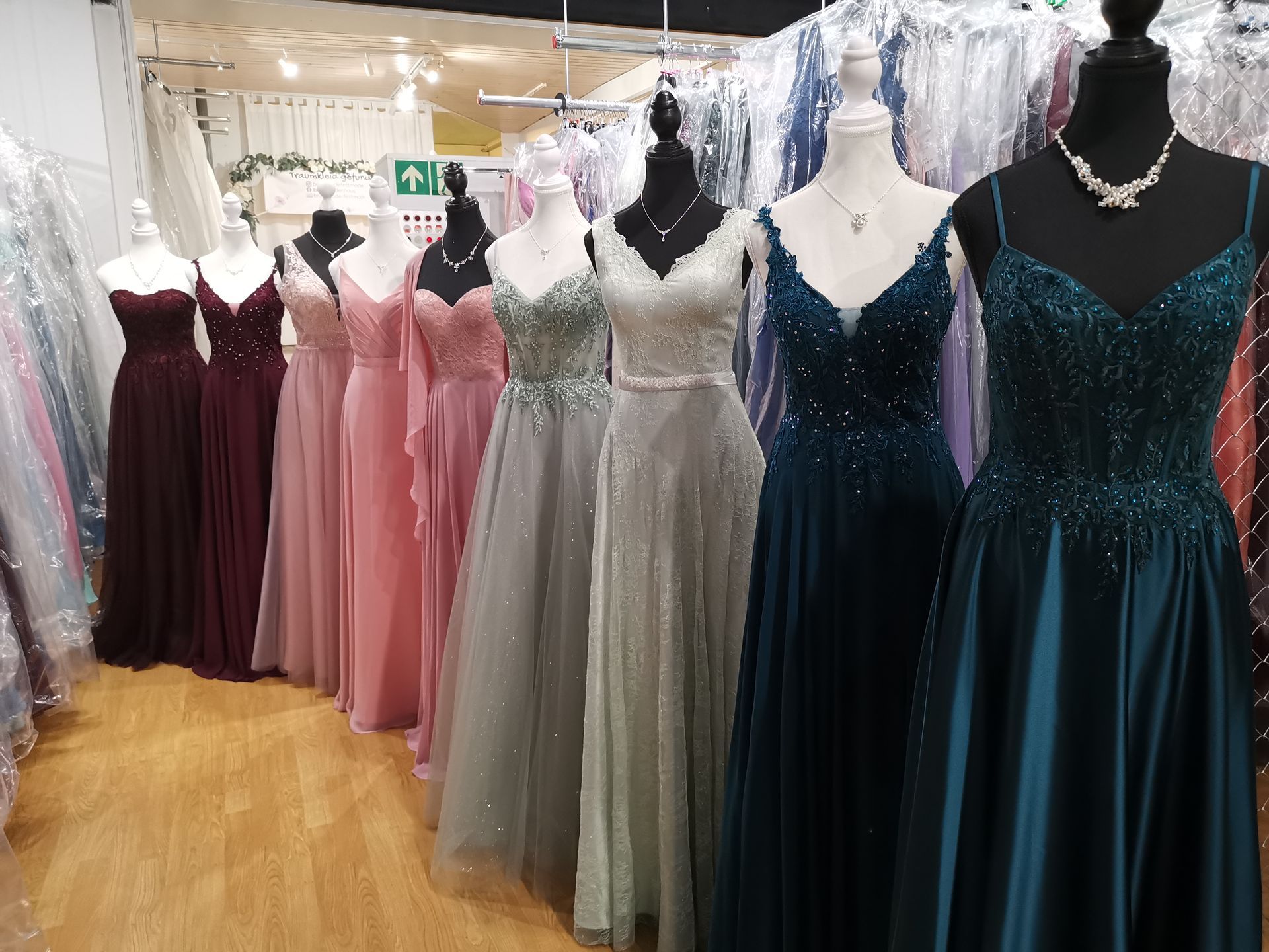 Festliche Kleider in einer Boutique, darunter paillettenbesetzte Kleider in Türkis, Schwarz, Grau, Rosa und Burgunderrot an Schaufensterpuppen.
