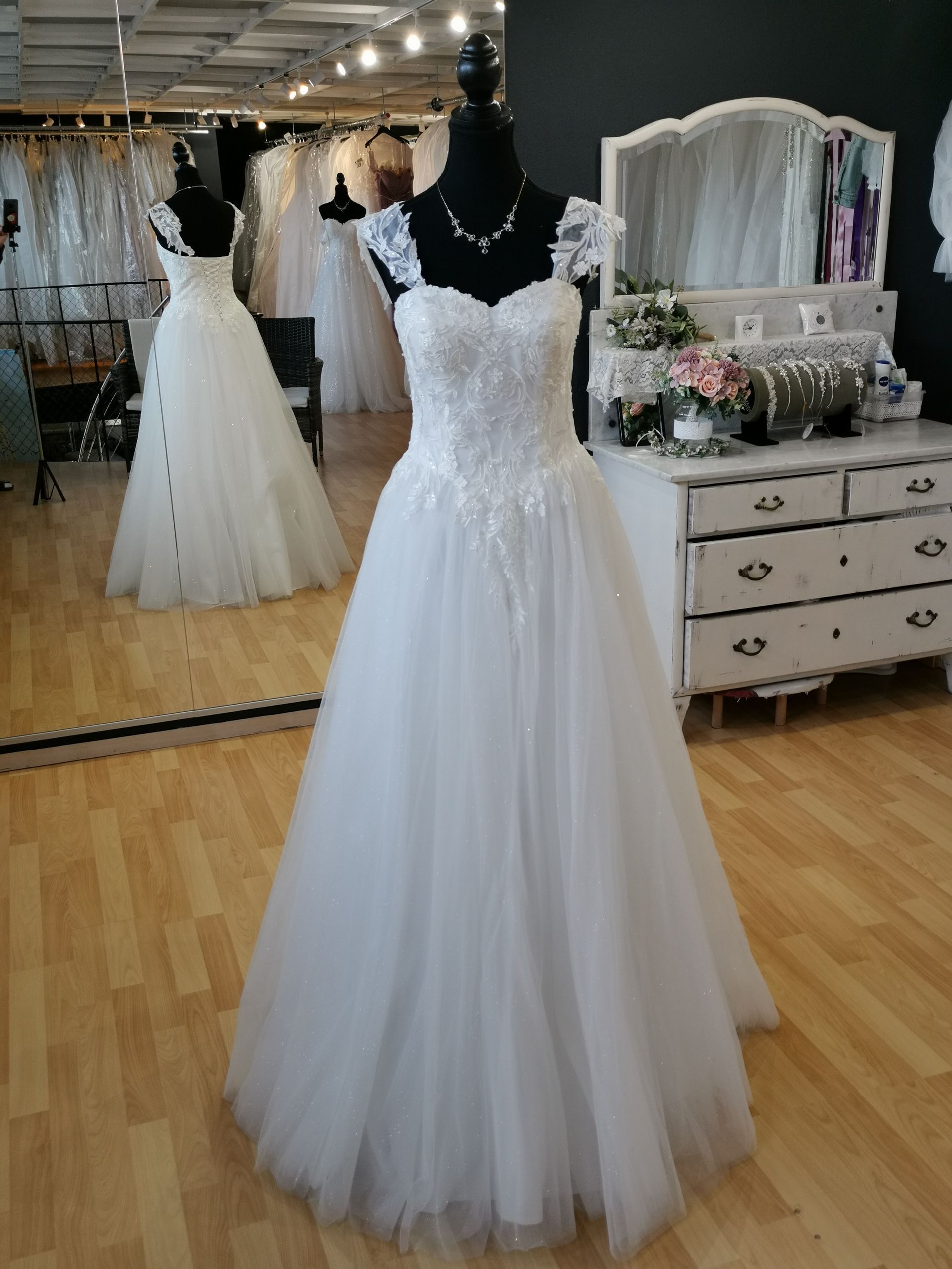 Weißes Spitzenhochzeitskleid auf einer schwarzen Schaufensterpuppe in einem Brautmodengeschäft mit Spiegeln und Kleidern im Hintergrund