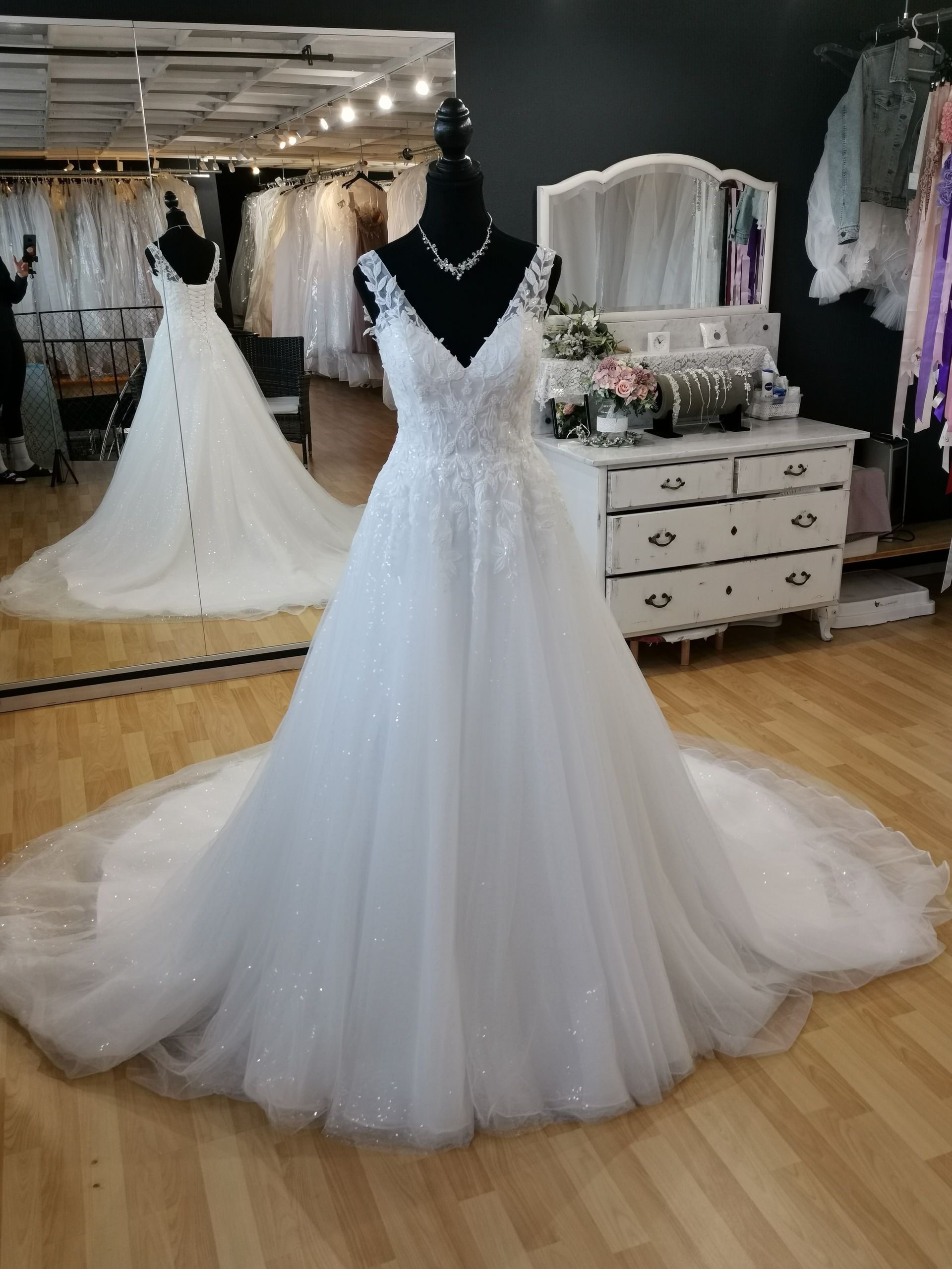 Weißes, perlenbesetztes Brautkleid an einer Schaufensterpuppe in einem Brautmodengeschäft mit verspiegelten Wänden und Kleiderständern.