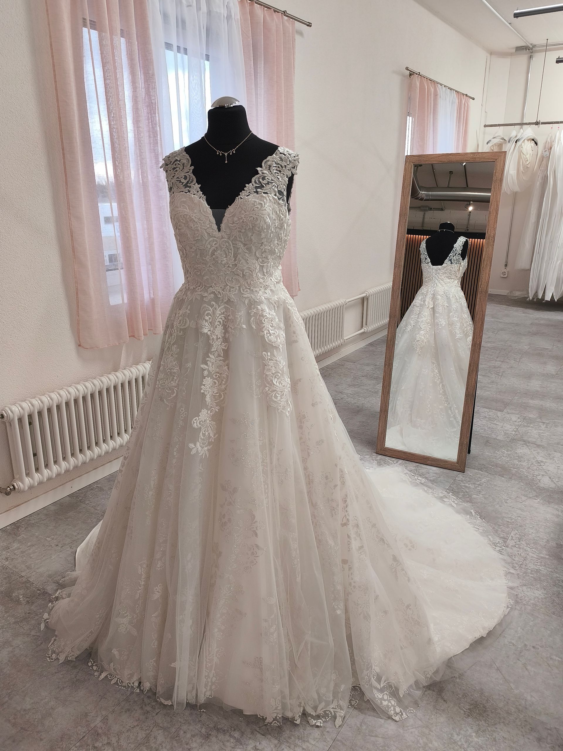Weißes Brautkleid aus Spitze an einer Schaufensterpuppe in einem hellen Brautmodengeschäft, mit einem Spiegel, der die Rückseite zeigt.
